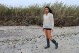 Andrelie standing in her custom hand embroidered jusi Barong Tagalog, black shorts and black boots on the sand with tall plants behind her at Dead Horse Bay Beach in Brooklyn, NY