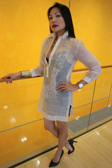 Anna stands in a dress length hand embroidered piña silk Barong Tagalog. Anna also wears a gold watch, gold armband and black high heel shoes. Anna stands with her left hand on her hip, right hand on a wood handrail with an orange wall behind her.