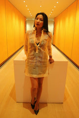 Anna stands and leans with hands resting on white table behind her in orange hallway. Anna wears a dress length hand embroidered piña silk Barong Tagalog