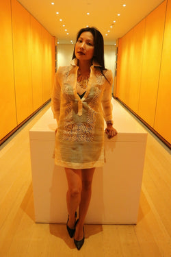 Anna stands and leans with hands resting on white table behind her in orange hallway. Anna wears a dress length hand embroidered piña silk Barong Tagalog