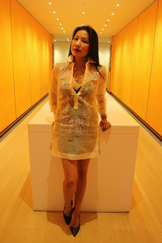 Anna stands and leans with hands resting on white table behind her in orange hallway. Anna wears a dress length hand embroidered piña silk Barong Tagalog