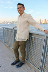 Angle side view of Arland standing and smiling in his hand embroidered piña silk Barong Tagalog, olive green pants and black shoes. Arland leans on grey gate with water and New York City skyline behind him.