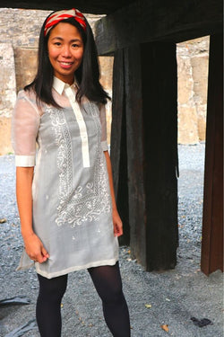 Bianca stands smiling in her dress length hand embroidered cocoon Barong Tagalog, black leggings and red and white headband next to dark wooden pillars and a stone wall behind her.