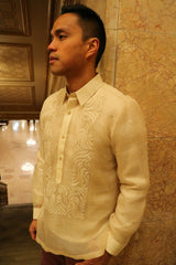 Side angled view of Blase from hips up wearing his hand embroidered piña silk Barong Tagalog with his hands at his sides, leaning his back on a marble wall