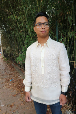 Shot of Bryan from his thighs up wearing his hand embroidered piña silk Barong Tagalog, blue jeans, black watch and glasses. Bryan stands on a brick walkway with dead leaves on the ground and bamboo branches and leaves behind him
