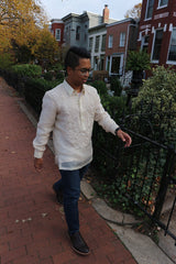 Bryan walks towards a wrought iron black gate with his left hand extended towards it. Bryan wears a hand embroidered piña silk Barong Tagalog, blue jeans, dark brown shoes, black watch and glasses. Also pictured is the sidewalk, shrubs, trees and a row of houses in the background  
