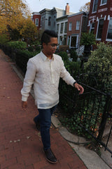 Bryan walks towards a wrought iron black gate with his left hand extended towards it. Bryan wears a hand embroidered piña silk Barong Tagalog, blue jeans, dark brown shoes, black watch and glasses. Also pictured is the sidewalk, shrubs, trees and a row of houses in the background