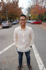 Bryan stands in the middle of the street smiling with his hands in his pockets, wearing his hand embroidered piña silk Barong Tagalog, blue jeans, dark brown dress shoes, glasses, black wrist watch. Also pictured is the road behind him, parked cars and trees