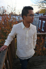 Bryan stands on a wooden deck terrace with his right hand on the wood railing, left hand in his pocket, looking to his left. Bryan wears a hand embroidered piña silk Barong Tagalog, blue jeans and glasses. There are tree top branches, leaves and the tops of houses behind him