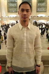 Chris pictured from the waist up with his hands at his sides. Chris wears his hand embroidered piña silk Barong Tagalog, dark pants and black bracelet. Behind Chris is Grand Central Terminal in NYC with crowds of people standing and walking