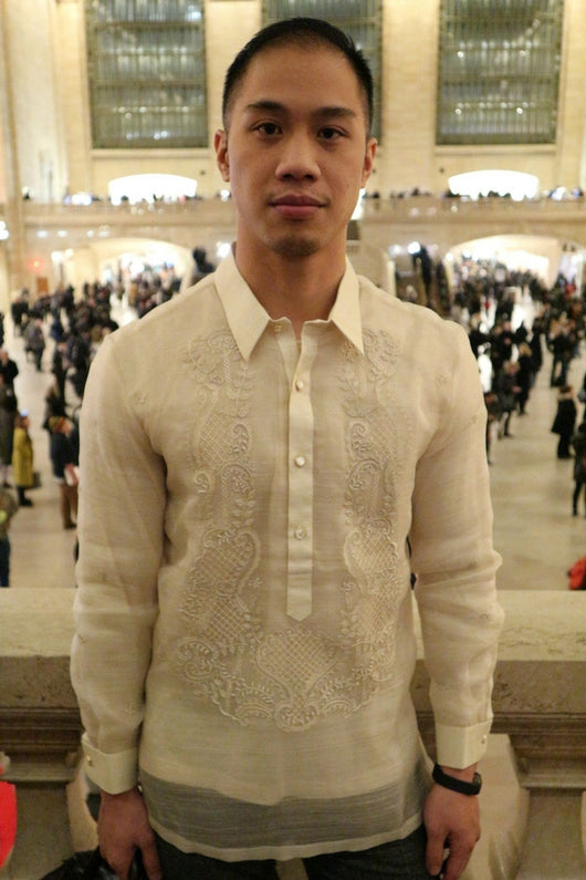 Chris pictured from the waist up with his hands at his sides. Chris wears his hand embroidered piña silk Barong Tagalog, dark pants and black bracelet. Behind Chris is Grand Central Terminal in NYC with crowds of people standing and walking