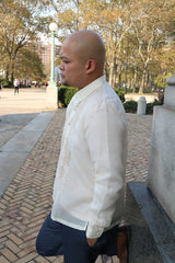 Side view of Chris U in Prospect Park, Brooklyn, in his hand embroidered jusi Barong Tagalog, blue slacks and brown shoes, leaning back on a marble and stone base with his hands in his pockets and his right leg bent and right foot pressed on marble base. Brick paved ground, street lamp and trees in background