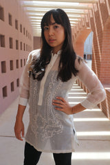 Coley from the knees up with her left hand on her hip and the right hand hanging at her side. Coley wears a dress length hand embroidered cocoon Barong Tagalog with a black tank top underneath that and black leggings. She is standing in a southwestern architecture style structure with sun rays shining in