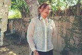 Iris B stands in front of a tree with wooden fence with green vines, and other trees behind them. Iris wears her blouse length full button down hand embroidered piña silk Barong Tagalog. She also wears black feather earrings, silver bracelets and black dress underneath her barong