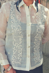 Product picture of the hand embroidered piña silk Iris B Barong Tagalog. The barong buttons all the way down. Model wears black feather earrings, a silver bracelet and black dress underneath her barong.