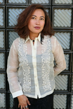 Iris B wears a full button down hand embroidered piña silk Barong Tagalog with black dress underneath. Iris stands in front of a wall with textured tiles