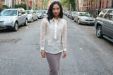 Jenny stands in the middle of the street with cars on both sides and surrounded by houses and apartment buildings. Jenny wears a hand embroidered piña silk Barong Tagalog, beige tank top under the barong, grey skirt, gold bracelets. Jenny looks to her left with her knee bent