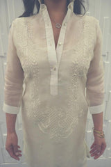 Product picture of the hand embroidered cocoon Jeri Barong Tagalog. Jeri wears a heart pendant necklace and brass bangles on her left wrist. Jeri stands in front of a textured white wall. 