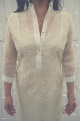 Product picture of the hand embroidered cocoon Jeri Barong Tagalog. Jeri wears a heart pendant necklace and brass bangles on her left wrist. Jeri stands in front of a textured white wall.