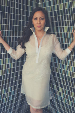 Jeri stands in the corner of two multicolor tile walls with her hands resting on the wall. Jeri wears her dress length hand embroidered cocoon Barong Tagalog, a beige minidress underneath her barong, hoop earrings, heart pendant necklace, gold bangles and diamond ring on her left arm and hand
