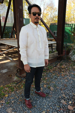 Angled shot of John-T standing on a gravel walkway in front of a wooden table and steel beams. John-T wears a hand embroidered jusi Barong Tagalog, dark sunglasses, dark blue jeans and dark red Doc Marten boots. There are trees and green buildings in the background