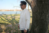 Side view of John-T in his hand embroidered jusi Barong Tagalog, dark sunglasses and dark blue jeans. There is a tree to the right of John-T. Grass, a gate, a body of water and buildings in the background