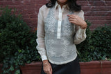 Product shot of the hand embroidered piña silk Kristina Barong Tagalog. Kristina wears a black tank top underneath her barong, black perforated leather miniskirt, gold bracelets on each wrist. There is a brick wall and shrubbery behind Kristina 