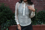 Product shot of the hand embroidered piña silk Kristina Barong Tagalog. Kristina wears a black tank top underneath her barong, black perforated leather miniskirt, gold bracelets on each wrist. There is a brick wall and shrubbery behind Kristina