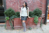 Kristina wears a hand embroidered piña silk Barong Tagalog, a black tank top underneath her barong, black perforated leather miniskirt, gold bracelets on each wrist and lavendar suede thigh high boots.. Kristina stands in front of a brown plant stand.  There is a brick wall and shrubbery behind Kristina 