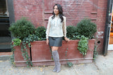 Kristina wears a hand embroidered piña silk Barong Tagalog, a black tank top underneath her barong, black perforated leather miniskirt, gold bracelets on each wrist and lavendar suede thigh high boots.. Kristina stands in front of a brown plant stand.  There is a brick wall and shrubbery behind Kristina
