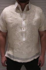 Product shot of the hand embroidered cocoon Lakhi Barong Tagalog with short sleeves. Lakhi wears black jeans. His hands are to his side. There is an off-white textured well behind him.