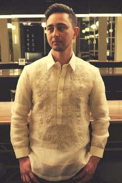 Half body photo of Mansour standing in his hand embroidered piña silk Barong Tagalog. Mansour stands in front of a bar and mirrors. His hands are over his front jeans pockets.