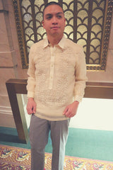 Mark leans back on a gold bar in front of a decorative window. There are marble walls and a green and red carpet. Mark wears his hand embroidered piña silk Barong Tagalog and grey slacks