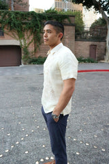 Side view of Matt standing in the middle of the street with his hands in his pockets in his short sleeve hand embroidered jusi Barong Tagalog. Matt also wears a chamisa de chino underneath his barong, blue pants and a wrist watch on his left wrist. There are metal studs in the street in a pattern. There is a brick garage covered in ivy, trees and other distant buildings in the background