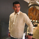 Michael stands in his hand embroidered cocoon Talisay Barong Tagalog. Michael wears a chamisa de chino underneath his barong, a black watch on his left wrist and a black beaded bracelet on his right wrist. He stands on a balcony with panes of protective glass behind him. There's a purple wall to the left. There are wooden organ pipes on the back wall. There are furniture, lamps, tables and rugs on the lower floor in the background