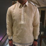 Product photo of the hand embroidered cocoon Talisay Barong Tagalog. Michael wears a chamisa de chino underneath his barong, a black watch on his left wrist and a black beaded bracelet on his right wrist. He stands on a balcony with panes of protective glass behind him. There's a purple wall to the left