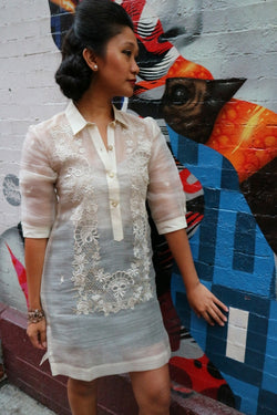 Rachelle stands in her dress length hand embroidered piña silk Barong Tagalog in front of an Audrey Hepburn mural in Little Italy, New York City. She wears a beige tank top and black skirt underneath her barong. She stands at an angle in front of the wall with her left hand touching it.