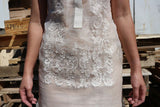 Close up on the bottom of the calado hand embroidery on the piña silk Rechelle Barong Tagalog. Rechelle wears a beige slip dress underneath her barong. There are stacked pallets and a white wall behind her.