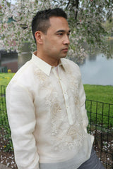 Angled view of Ricky standing in Central Park. Ricky wears a hand embroidered piña silk Barong Tagalog and grey pants. He wears a chamisa de chino underneath his barong. There is a gate, grass, a pond and cherry blossom tree behind him 