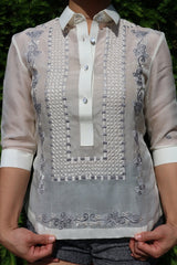 Product picture of the jusi Rissa Barong Tagalog. Rissa pulls down on the bottom of the barong with her hands. She also wears grew shorts. There is green pine in the background