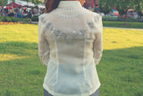 Product shot of the back embroidery of the jusi Rissa Barong Tagalog. There are grass, trees, vendor booths and vehicles in the background