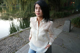 Robin sits on a concrete barrier in her hand embroidered piña silk Barong Tagalog, white tank top underneath her barong, and blue jeans. Robin's hands hold the bottom of her barong. Concrete and stone walkways, a pond and weeping willow tree are in the background