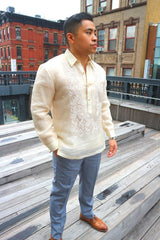 Rodrigo stands in his hand embroidered piña silk Barong Tagalog, chamisa de chino under his barong, grey and blue plaid slacks, and light brown dress shoes. Photo shows his full body standing at an angle with his right hand in his pocket, his left hand hanging to his side, and looking to his left. Rodrigo stands at the High Line Park in New York City with stairs, railings and buildings behind him.
