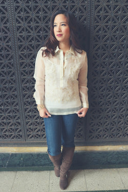 Roe stands in front of a decorated iron vent in her hand embroidered piña silk Barong Tagalog, white tank top underneath her barong, blue jeans, knee high brown boots and gold bracelet on her right hand. Roe stands with her back to the vent and looks to her right. Her hands hold the bottom of her barong