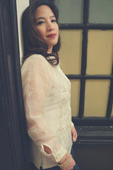 Side view of Roe in her hand embroidered piña silk Barong Tagalog. She wears a white tank top underneath her barong. She also wears a gold bracelet on her right wrist. There is a window with the glass panes painted yellow and green behind Roe. Roe looks into the camera.
