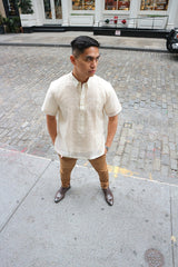 Downward view of Ryan standing on the sidewalk with a cobblestone road, the opposite sidewalk and a white building with windows behind him. Ryan wears a short sleeve hand embroidered jusi Barong Tagalog, a chamisa de chino underneath his barong, brown slacks, dark brown shoes and a silver watch on his left wrist. Ryan stands with his hands in his pockets and he looks to his left