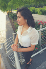 Side view of Ryann standing with her hands resting in front of her on guardrails in a park in Georgetown. Ryann wears her jusi Barong Tagalog tucked into her black skirt. She wears a white tank top underneath her barong. She also wears open toe black shoes. There are flowers, trees, a paved walkway and other plants in the background. Ryann looks out towards the water