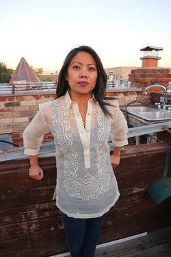 Sarahlynn stands in her hand embroidered piña silk Barong Tagalog, black tank top underneath her barong, and dark blue jeans. She stands leaning back with her hands resting on a wooden wall. She is on a rooftop, and a brick wall, sink, chimney, other rooftops, trees and the sky is in the background