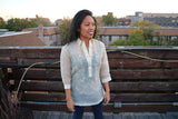 Sarahlynn stands on a wooden rooftop deck with a wooden wall, buildings, trees and the sky in the background. Sarahlynn wears a hand embroidered piña silk Barong Tagalog, black tank top underneath her barong and dark blue jeans. She stands with her hands at her sides looking to her left and smiling