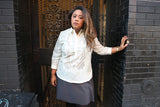 Stephanie stands in a black doorway with her left hand pressed against the wall next to her. She wears a hand embroidered jusi Barong Tagalog, black 3/4 sleeve shirt underneather her barong and grey skirt.